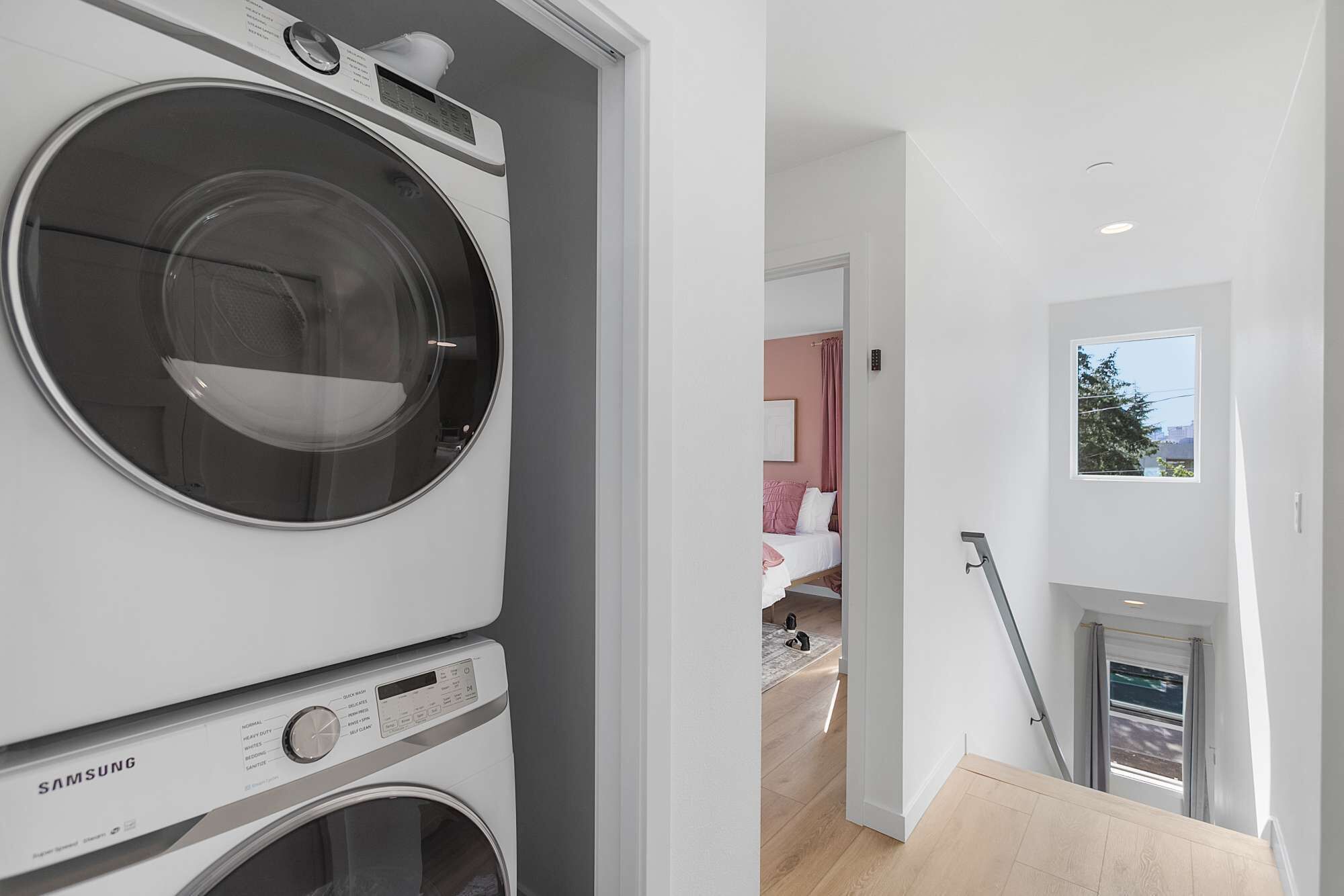 A modern laundry room with a Samsung washer and dryer, a hallway leading to a bedroom with a bed, and a window with a view.
