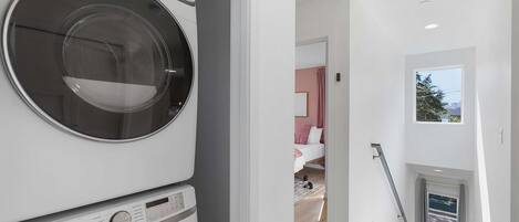A modern laundry room with a Samsung washer and dryer, a hallway leading to a bedroom with a bed, and a window with a view.