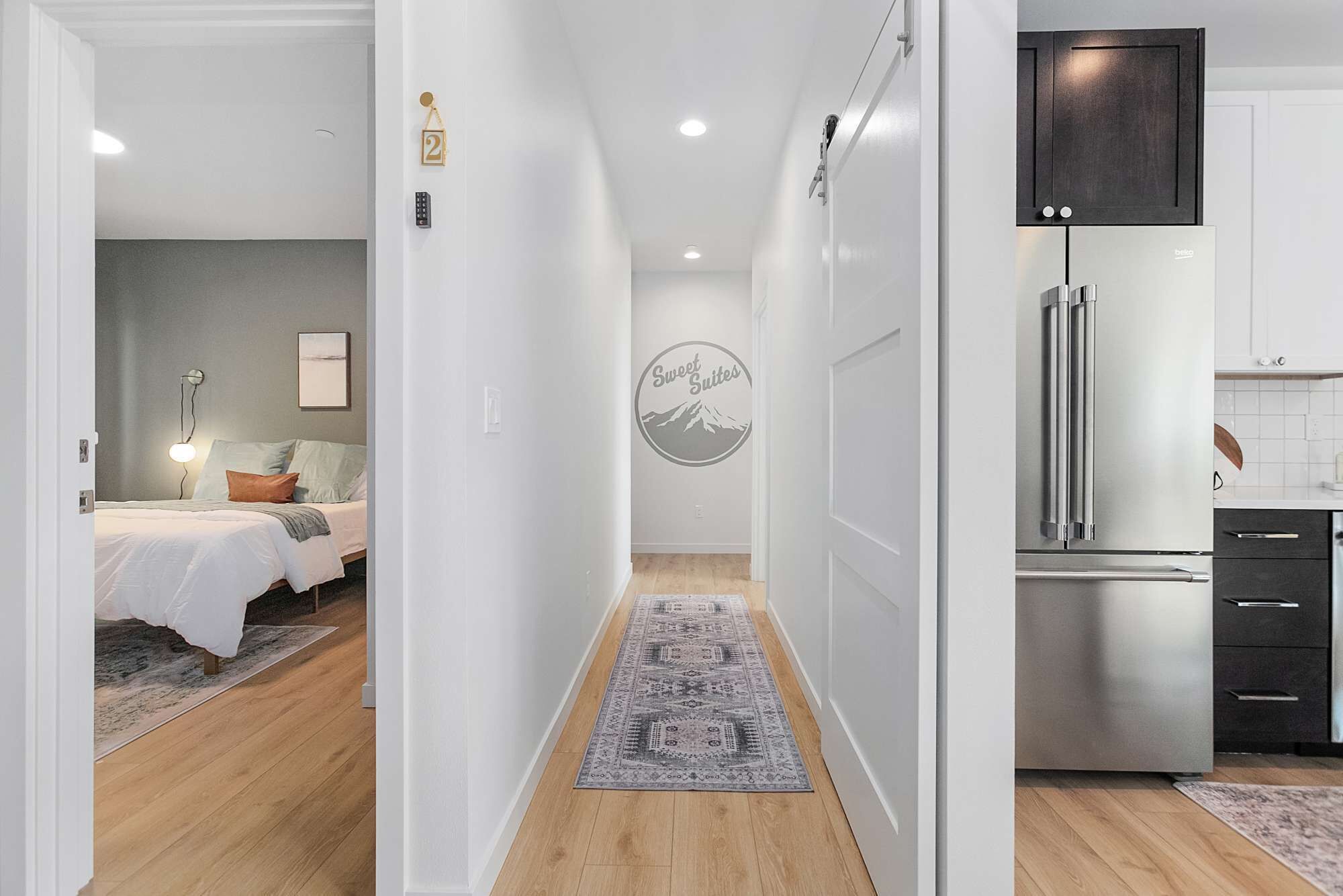 Una habitación de hotel moderna con cama, mesita de noche y lámpara fijada a la pared. Un pasillo conduce a una zona de cocina con un refrigerador de acero inoxidable y armarios.