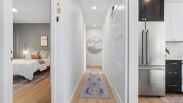 Una habitación de hotel moderna con cama, mesita de noche y lámpara fijada a la pared. Un pasillo conduce a una zona de cocina con un refrigerador de acero inoxidable y armarios.