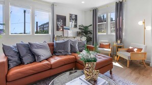 A modern living room with a brown leather sofa, a glass coffee table, and a white armchair. There are large windows with curtains, a bookshelf, and a plant. - Kenny's Sweet Suite - 84 Walkscore!! (Seattle)