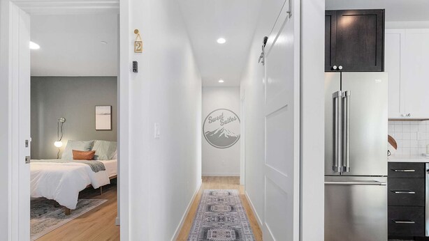 A modern hotel room with a bed, bedside table, and a wall-mounted light fixture. A hallway leads to a kitchen area with a stainless steel refrigerator and cabinets. - Kurt's Sweet Suite - 84 Walkscore!! (Seattle)