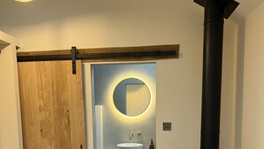 Une salle de bain moderne avec une porte en bois, un miroir rond, un lavabo blanc et un poêle à bois noir.
