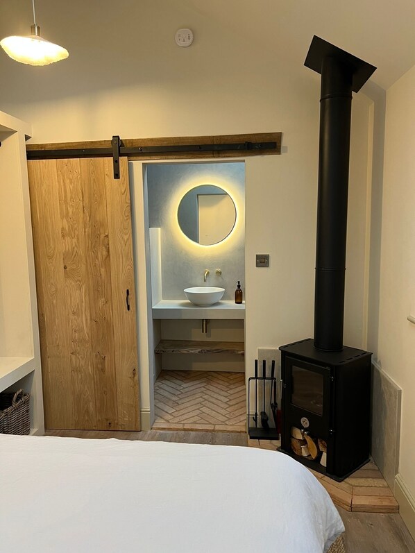 Een moderne badkamer met een houten deur, een ronde spiegel, een witte wastafel en een zwarte houtkachel.