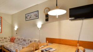Een gezellige kamer met een houten eettafel, een bank met decoratieve kussens, een aan de muur bevestigde televisie en twee wandlampen.