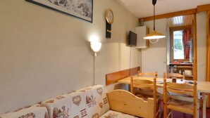 Een gezellige kamer met een houten bank, een eettafel met stoelen, een wandgemonteerde televisie en een lijst met een berglandschap.