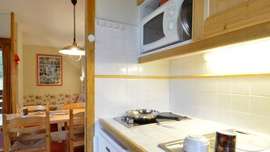 Een kleine keuken met een magnetron, een fornuis en een spoelbak. Er is een eettafel met stoelen en een hangende lamp.