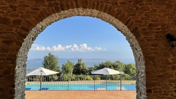 Um arco de pedra emoldura uma área de piscina com dois guarda-sóis e vista para o mar e montanhas.