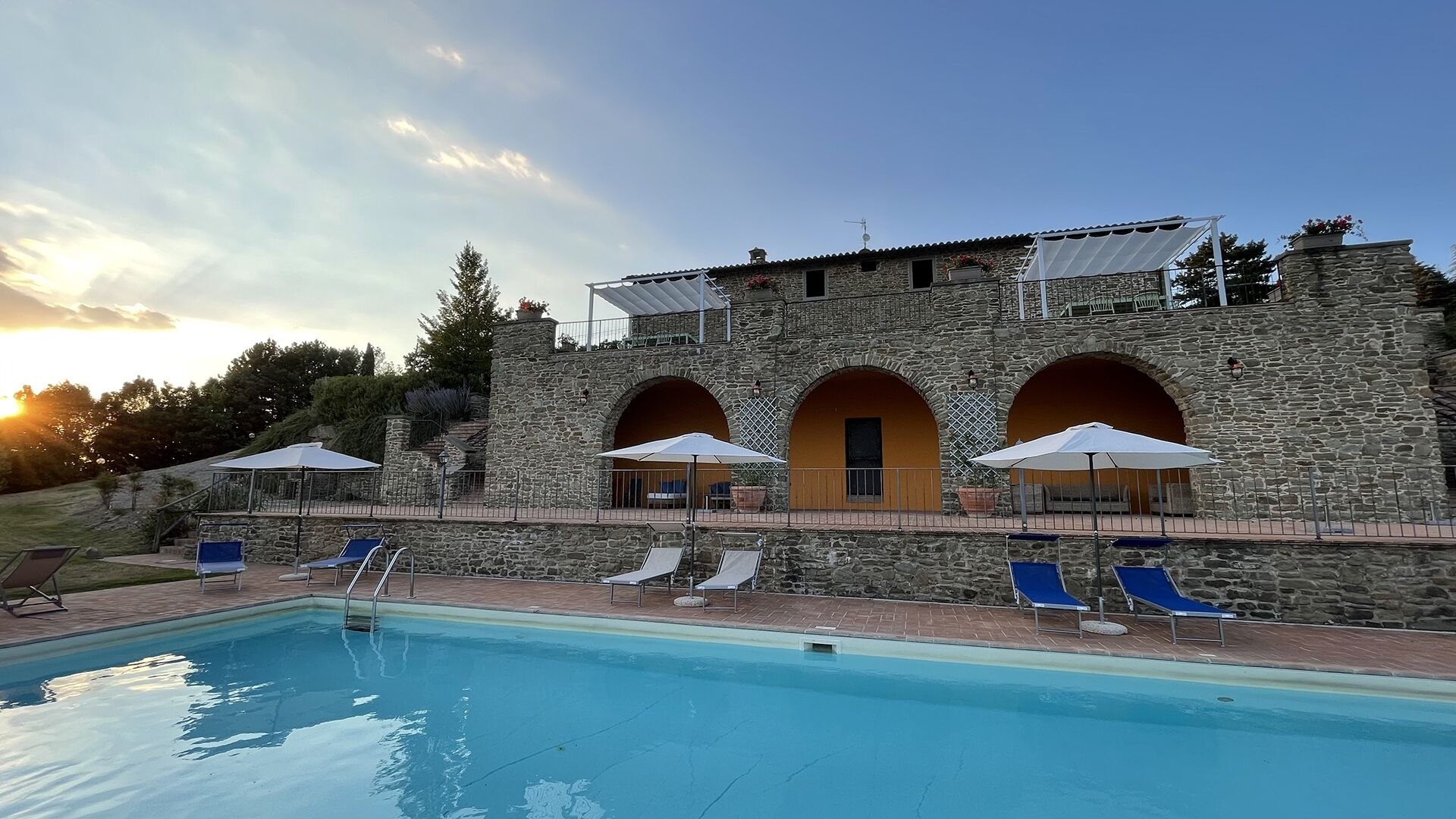 A stone building with a swimming pool, outdoor seating, and a sunset in the background.