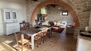 Uma cozinha rústica com parede de pedra, vigas de madeira, mesa de jantar com cadeiras, armário branco e um espaço de estar acolhedor com sofá e poltrona.