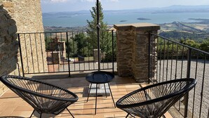 Uma varanda com duas cadeiras, uma mesa e um guarda-sol verde. Oferece vista para uma paisagem deslumbrante, com um corpo d'água e colinas ao longe.