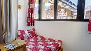 A small, single-bed room with a window overlooking a snowy mountain and buildings. There is a wooden nightstand with a glass of water and a newspaper. - Renovated 2-room apartment, ski-in/ski-out, 4 pers, WiFi (Les Menuires)