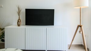 Una sala de estar moderna con un mueble blanco, un televisor de pantalla plana, una lámpara de pie y un florero con plantas secas.