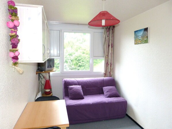 A small room with a purple sofa, a wooden table, a red hanging lamp, and a window with curtains. - Functional studio at the foot of the slopes with pool access (Le Corbier)