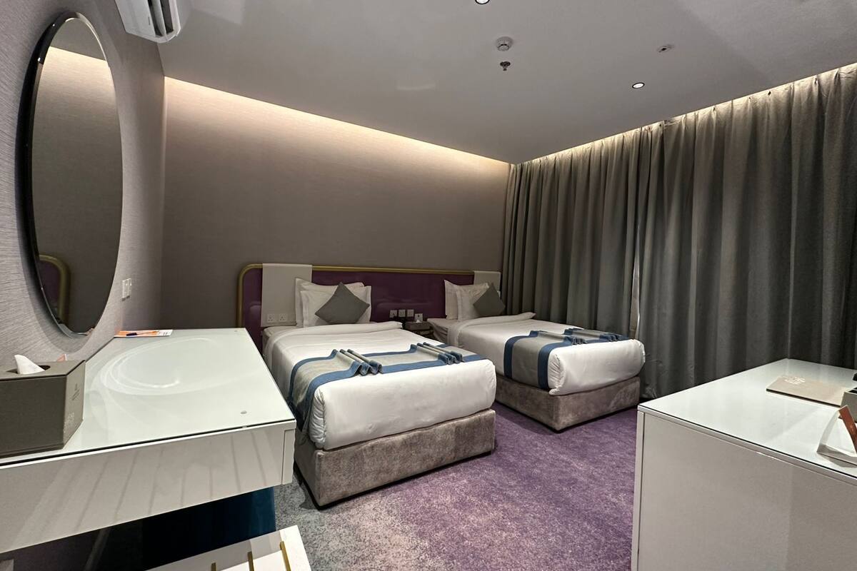 A hotel room with two beds, a large mirror, a vanity with a sink, and a window with curtains.