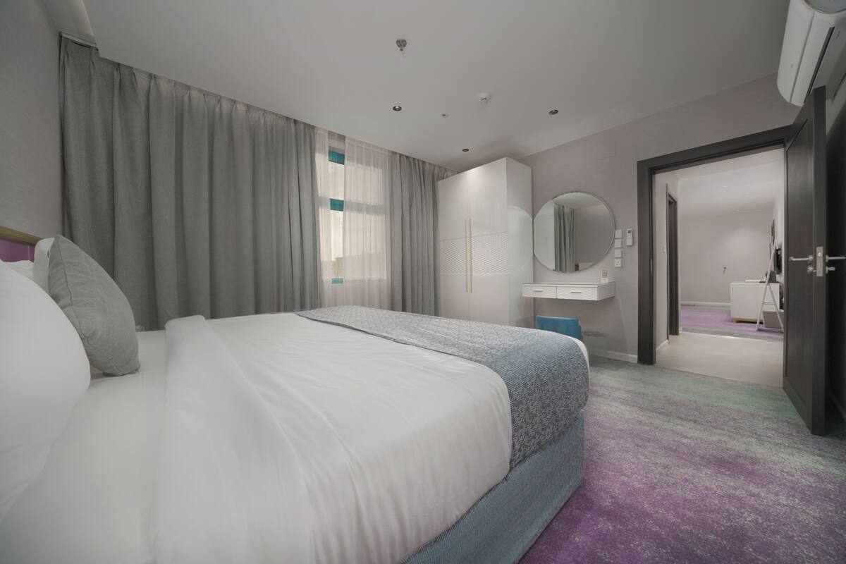 A modern hotel room with a large bed, a wardrobe, a mirror, and a bathroom visible through an open door.