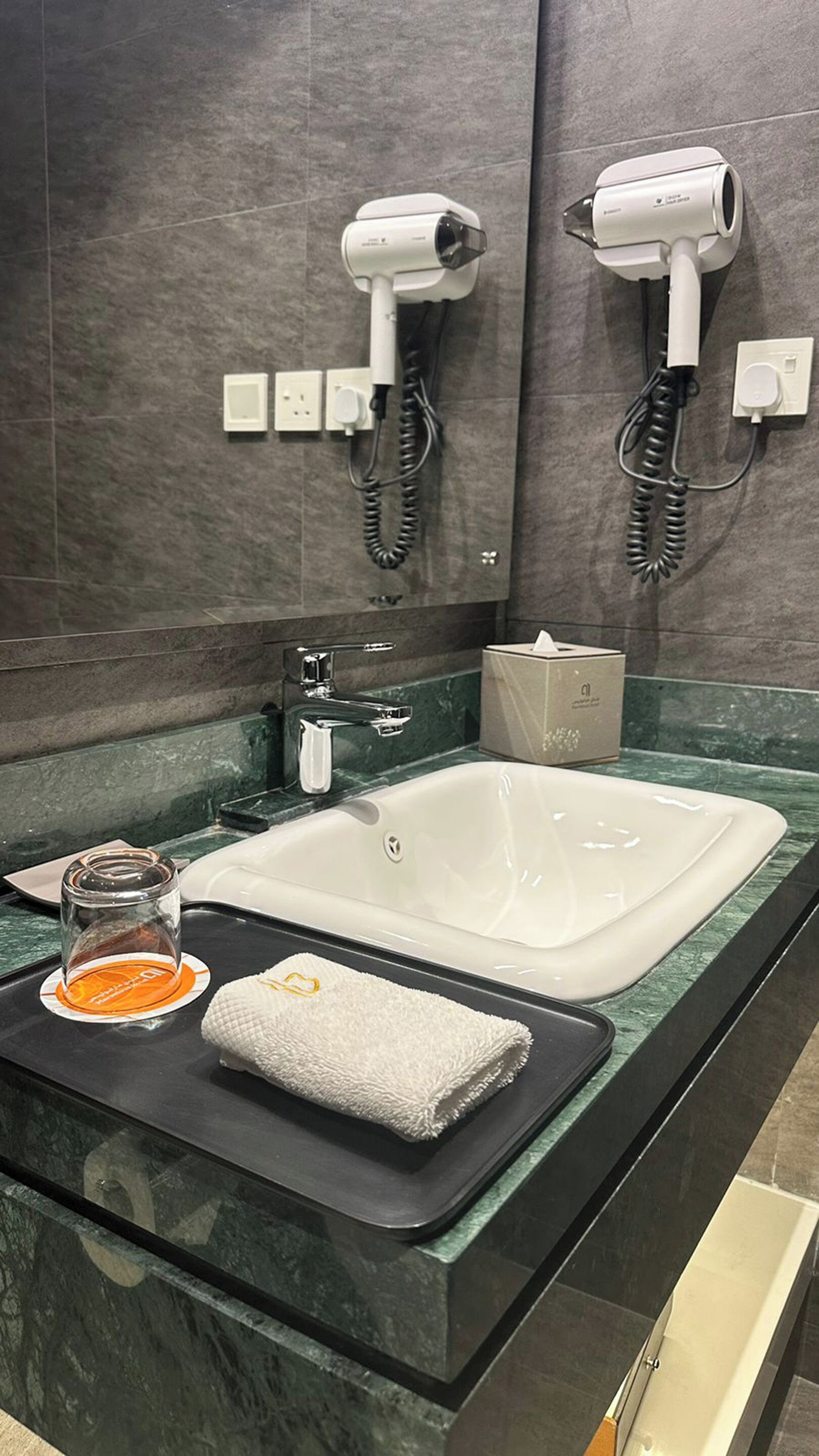 A bathroom with a large sink, two hair dryers mounted on the wall, a glass of orange juice, a towel, and a box of tissues on the counter.