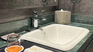 A bathroom with a large sink, two hair dryers mounted on the wall, a glass of orange juice, a towel, and a box of tissues on the counter.