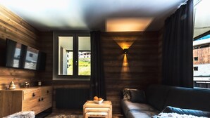 Un salón acogedor con suelo de madera, un sofá, una mesita con fruta, un televisor en la pared y una ventana con vistas a zonas verdes.