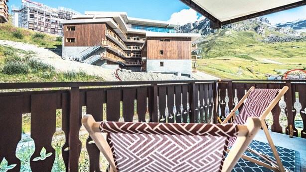 Un balcón con barandillas de madera y dos sillas con diseños, con vistas a un paisaje montañoso y un edificio moderno con elementos de madera y vidrio.