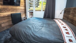 Un dormitorio con cama, televisor montado en la pared, ventana con cortinas y vista a edificios.
