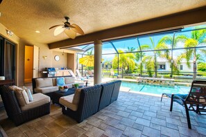 Zona junto a la piscina con mobiliario de mimbre, ventilador de techo y vistas a palmeras y a la piscina.