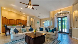 A spacious living room with a dark wooden coffee table, two white sofas with blue and green cushions, a patterned rug, and a ceiling fan. - Villa SunnySide- perfect for families and boaters (Cape Coral)