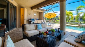 A spacious patio area with a sofa, wicker coffee table with fruit, and a view of a swimming pool and greenery. - Villa SunnySide- perfect for families and boaters (Cape Coral)