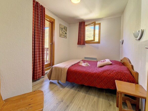 Une chambre à coucher avec un lit, une table de chevet en bois, une fenêtre avec des rideaux et un luminaire fixé au mur.