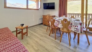 A dining area with a table set for four, wooden chairs, a television on the wall, and large windows with a view of the mountains. - Spacious apartment in Les Menuires, 3 rooms, at the foot of the slopes (Les Menuires)