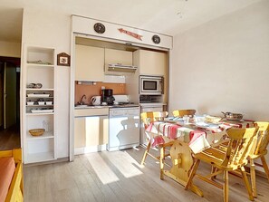 Cozinha compacta com micro-ondas, fogão e máquina de café. Há uma mesa de jantar com cadeiras e um pequeno armário com louças.