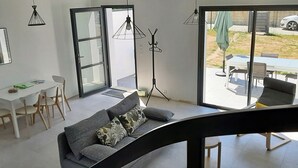 Ett modernt vardagsrum med en soffa, ett matbord och stolar. Det finns en stor skjutdörr i glas som leder ut till ett utomhusområde med ett bord och stolar.