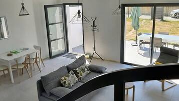 Un salon moderne comprenant un canapé, une table à manger et des chaises. Une grande porte-fenêtre coulissante donne accès à un espace extérieur aménagé avec une table et des chaises.