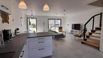 Une cuisine moderne dotée d’un îlot central, de meubles blancs et d’un éclairage suspendu. On y trouve un escalier avec des marches en bois et une rampe noire.
