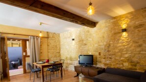 A cozy living room with a stone wall, a wooden ceiling, a television, a sofa, and a dining area with chairs and a table. - Cozy house in Daglan with fireplace and terrace (Daglan)