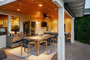 An outdoor patio with a wooden table and chairs, a ceiling fan, and a TV mounted on the wall.