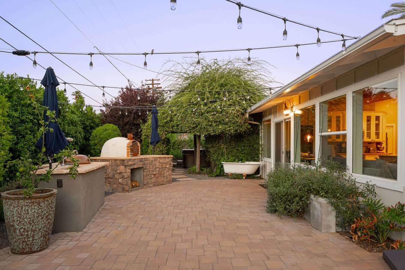 A patio with a stone barbecue, string lights, and a large window overlooking the garden.