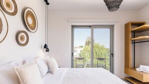 Un dormitorio con cama, almohadas, lámpara colgante, estante de madera y puerta corrediza de vidrio que da a un espacio exterior con mesa y sillas.