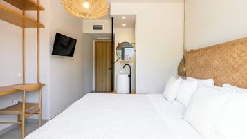 Ett modernt hotellrum med en stor säng, en tv som är monterad på väggen, en hylla i trä, en lampa av rotting och en spegel.