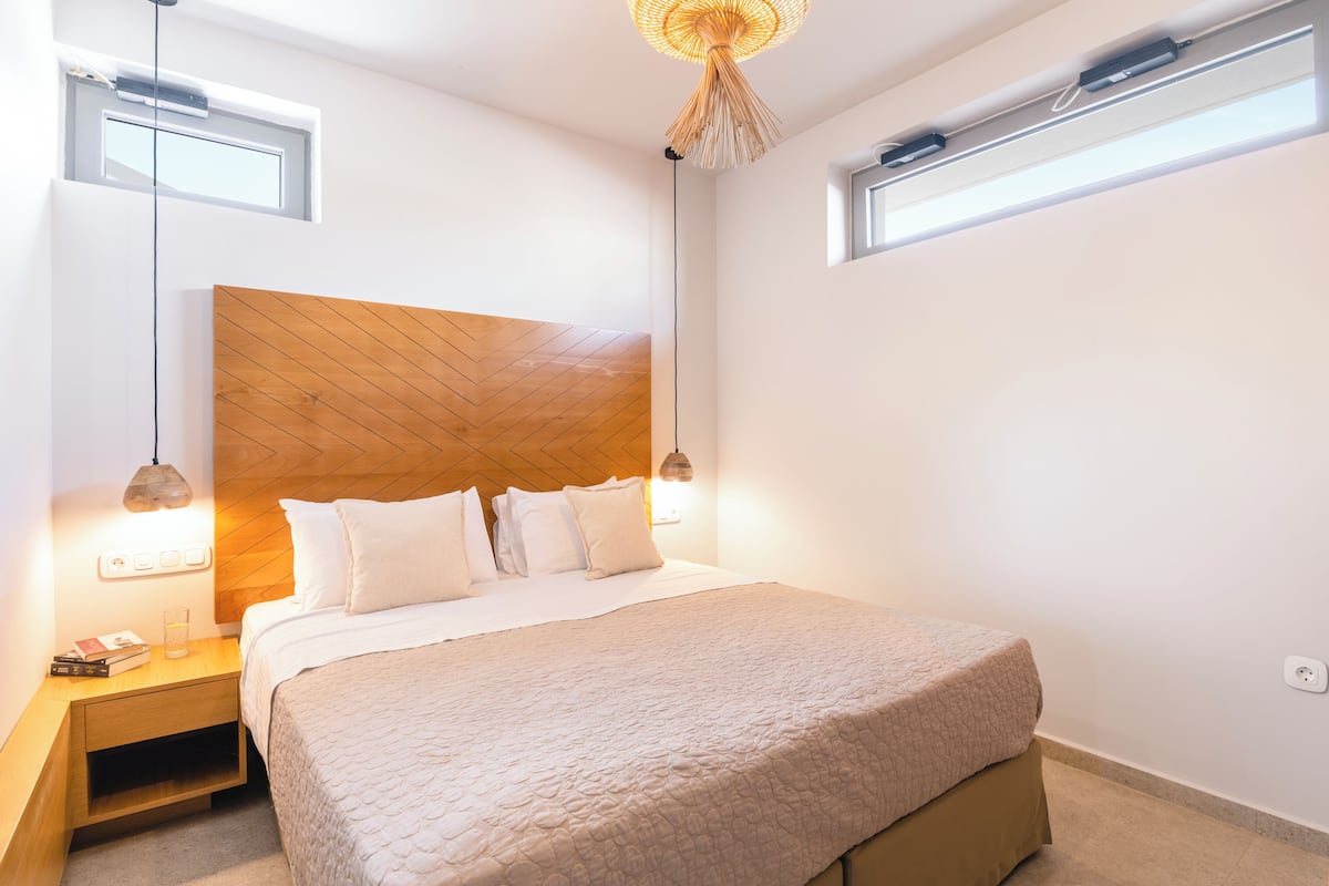 Un dormitorio con cabecera de madera, cama con ropa de cama blanca, lámparas de noche, mesita de noche y una ventana.