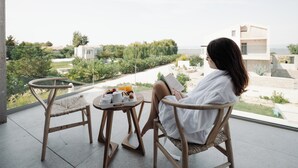 Una persona sentada en una silla de balcón leyendo un libro, con una bandeja de comida y bebidas sobre una mesita.