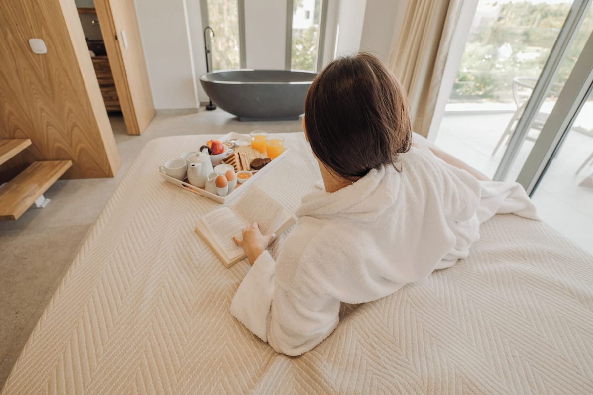 Una persona leyendo un libro en una cama, con una bandeja de desayuno y una bañera al fondo.