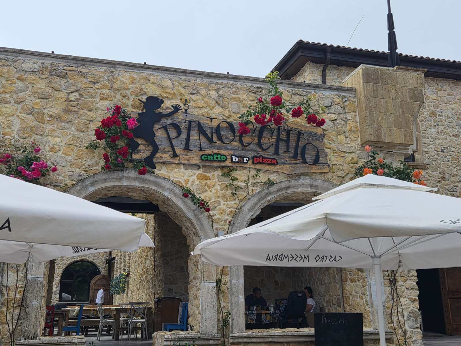 Ein Steinhaus mit einem Schild, auf dem 'Pinocchio' und 'caffe bar pizza' steht. Es gibt Tische und Sonnenschirme im Außenbereich.