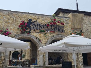 En byggnad i sten med en skylt där det står ”Pinocchio” och ”café, bar, pizzeria”. Det finns bord och parasoller utomhus.