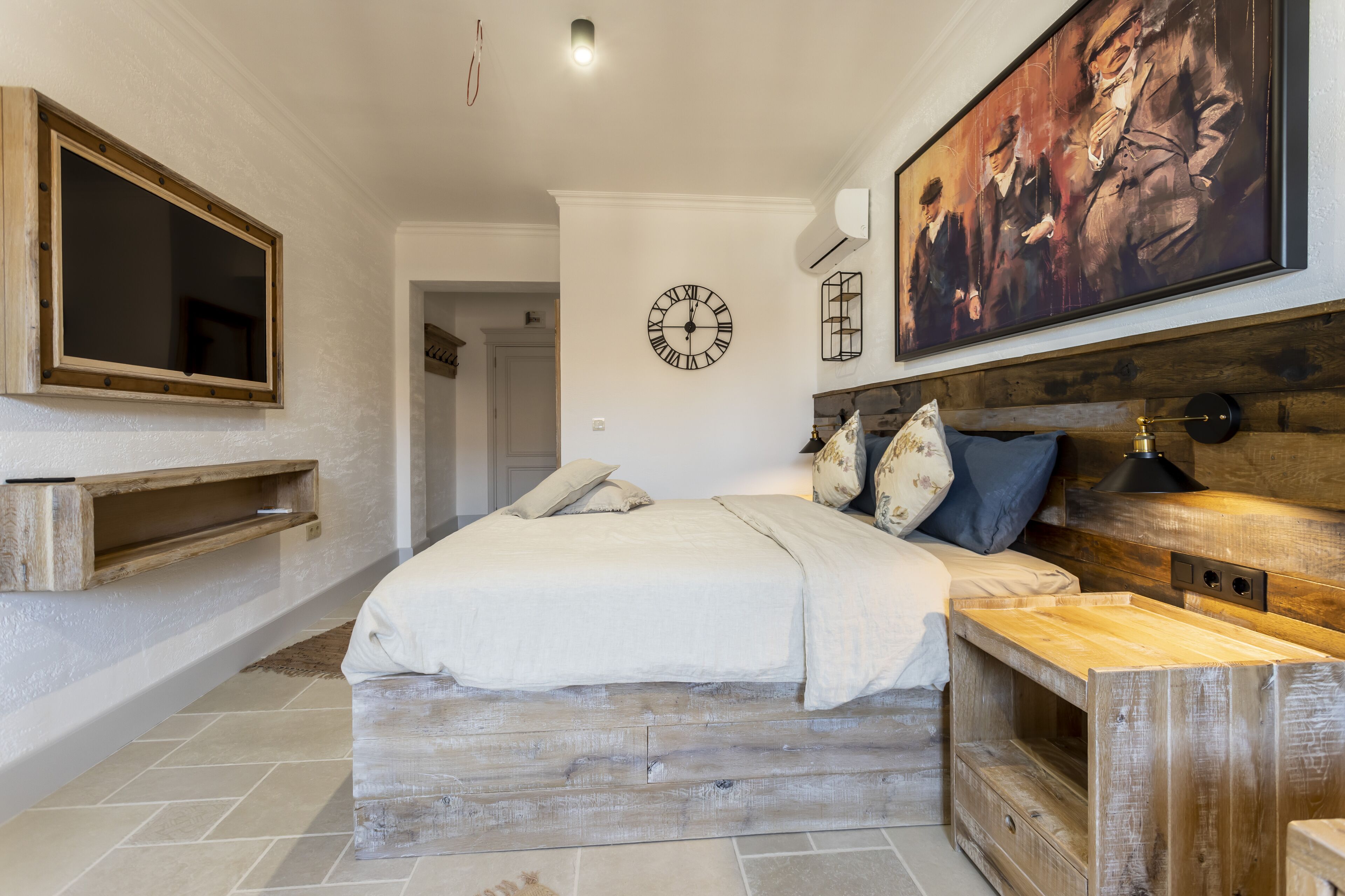 A bedroom with a large bed, wooden headboard, a mounted clock, a painting, and a TV on the wall.