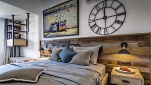 Ein modernes Schlafzimmer mit einem großen Bett, einem hölzernen Kopfteil, einer Wanduhr, einem Gemälde und einem Nachttisch mit einer Fruchtschale.