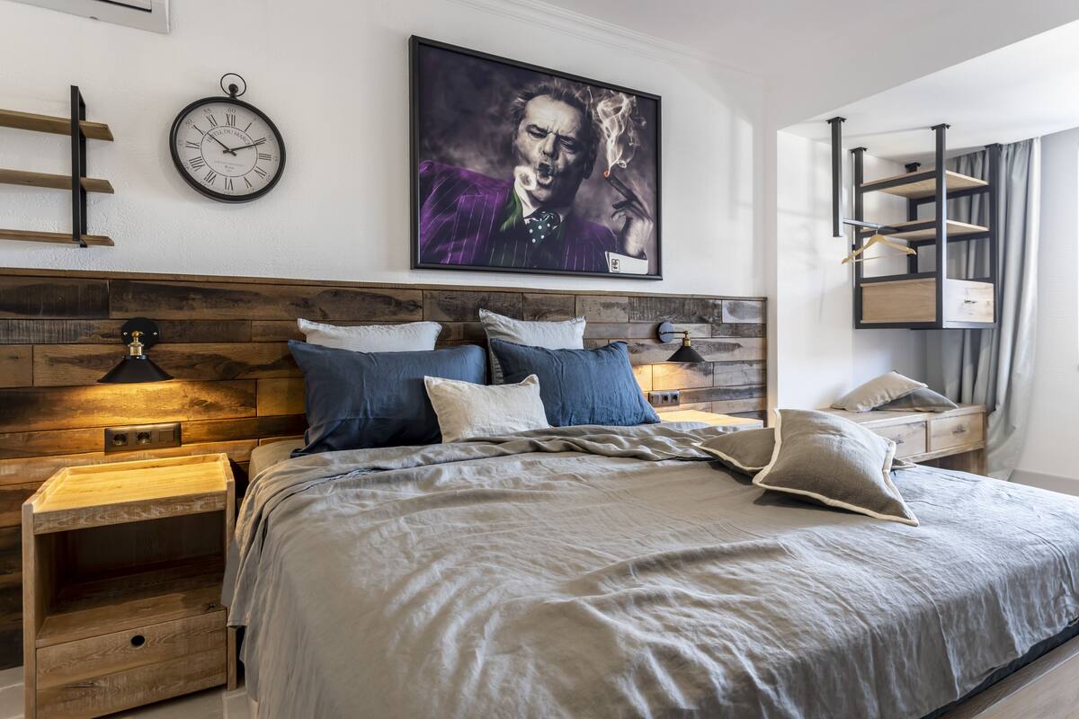 A modern bedroom with a wooden headboard, a large bed with grey bedding, a wall clock, and a framed artwork.