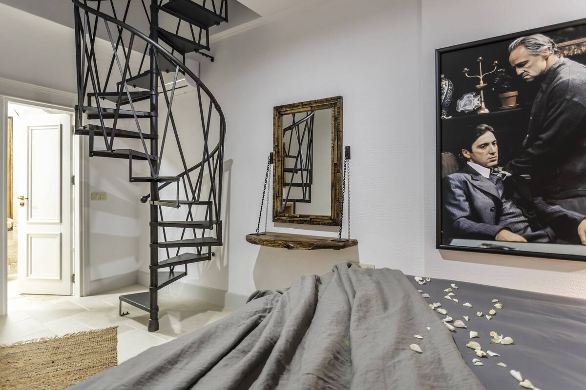 A bedroom with a spiral staircase, a bed with a gray blanket, a mirror, and a framed picture on the wall.
