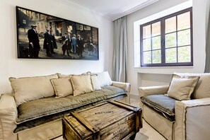 Ett vardagsrum med en beige soffa, en mindre fåtölj och ett soffbord med en klassisk design. En stor målning som föreställer en historisk scen hänger på väggen.
