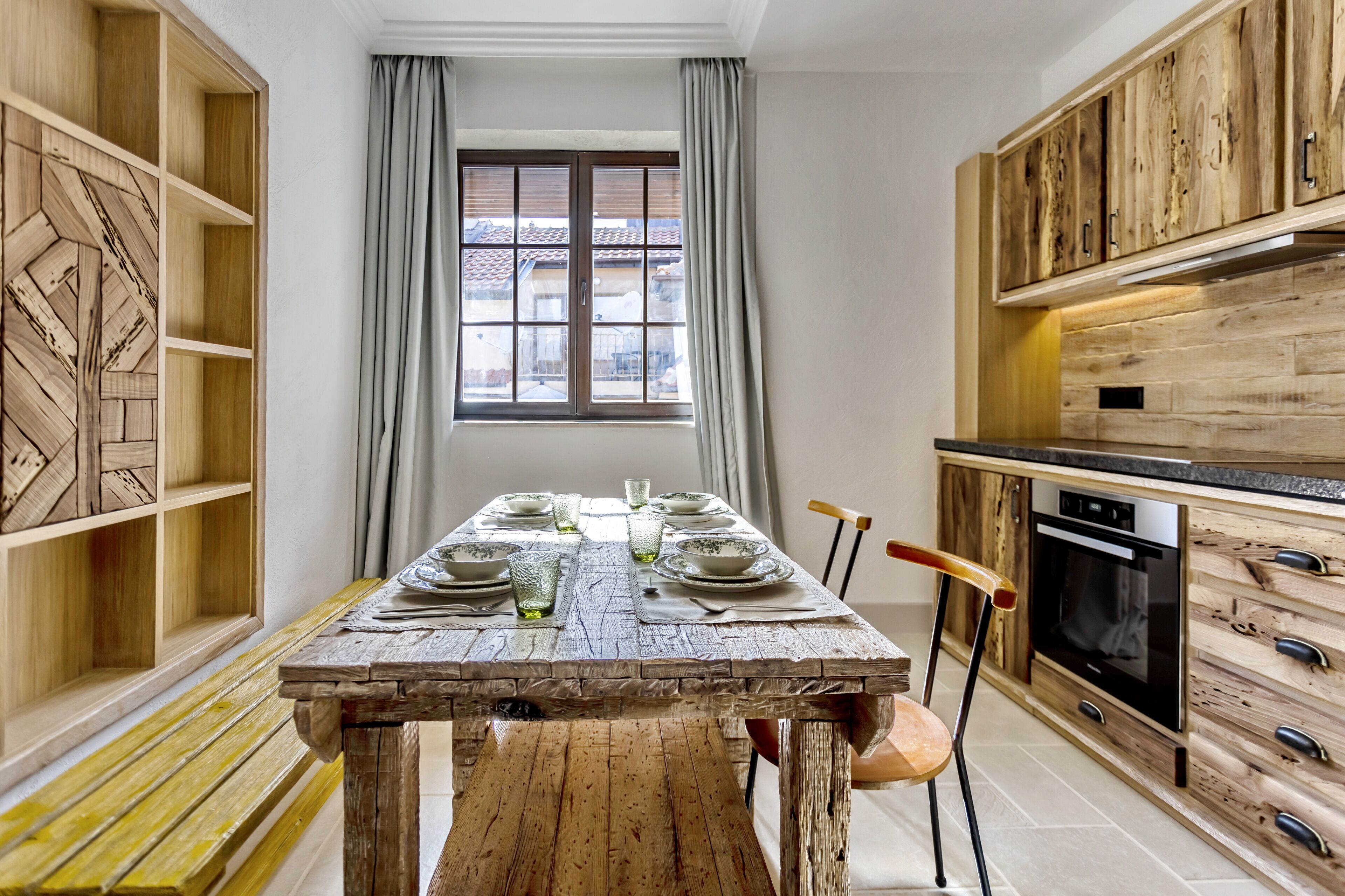 A kitchen with wooden cabinets, a dining table set with plates and glasses, and a window with curtains.